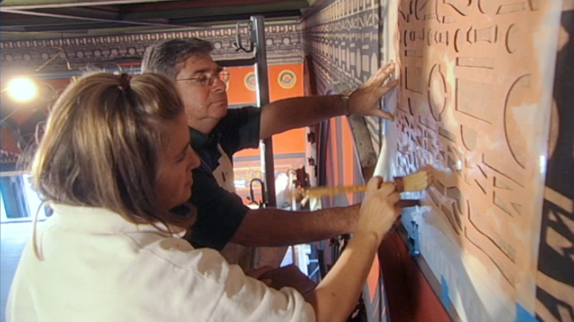 An older man and young woman work side by side in scaffolding near the ceiling of an historic church brushing tan-colored paint onto a hand-cut stencil of decorative geometric motifs that is taped to an interior wall; the bright orange colors of other parts of the church’s walls are in the background.