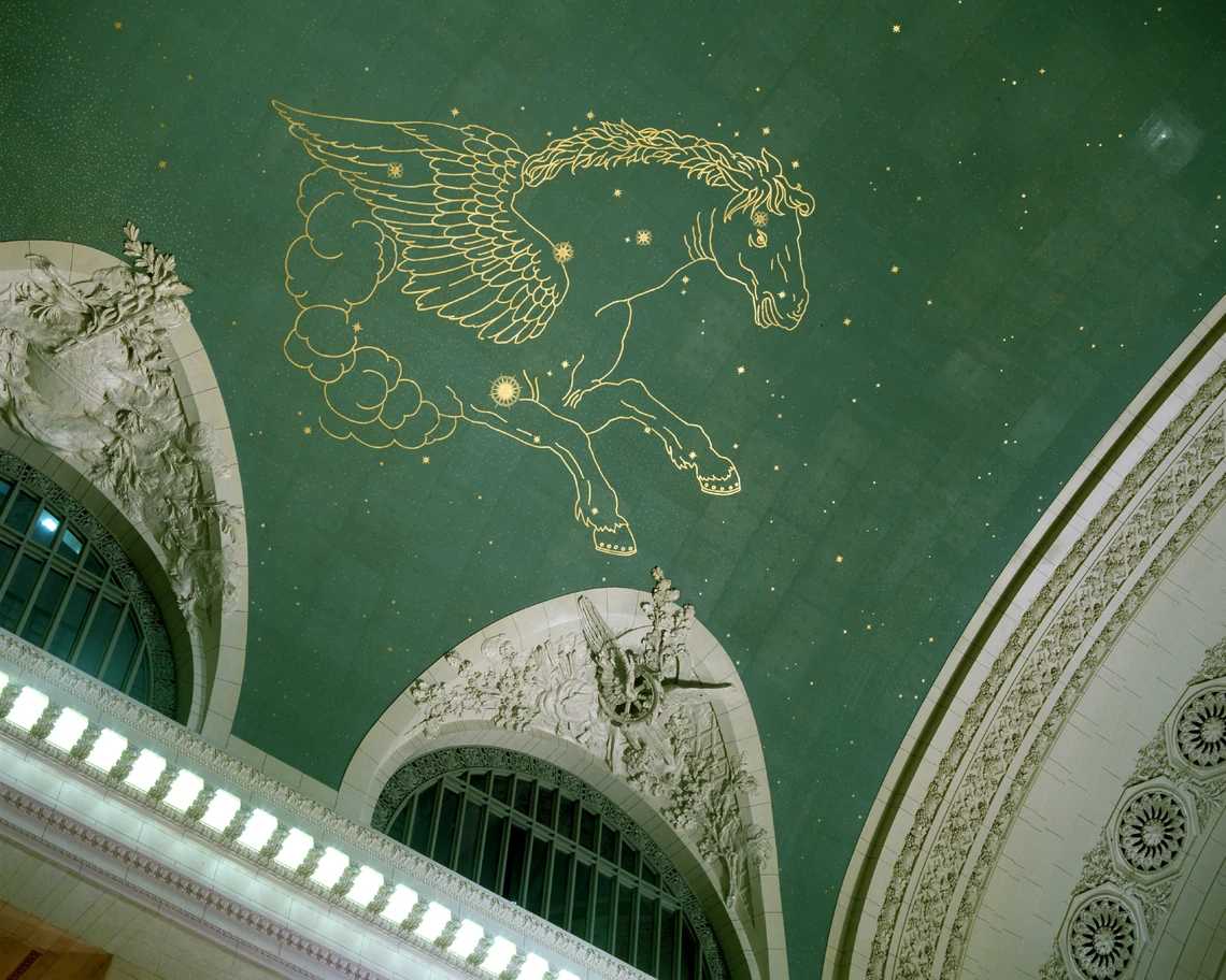 This partial image of Grand Central Terminal’s green star-studded ceiling shows the gold outlines of a galloping horse with wings; several carved ornamental arches are visible along the bottom of the ceiling.