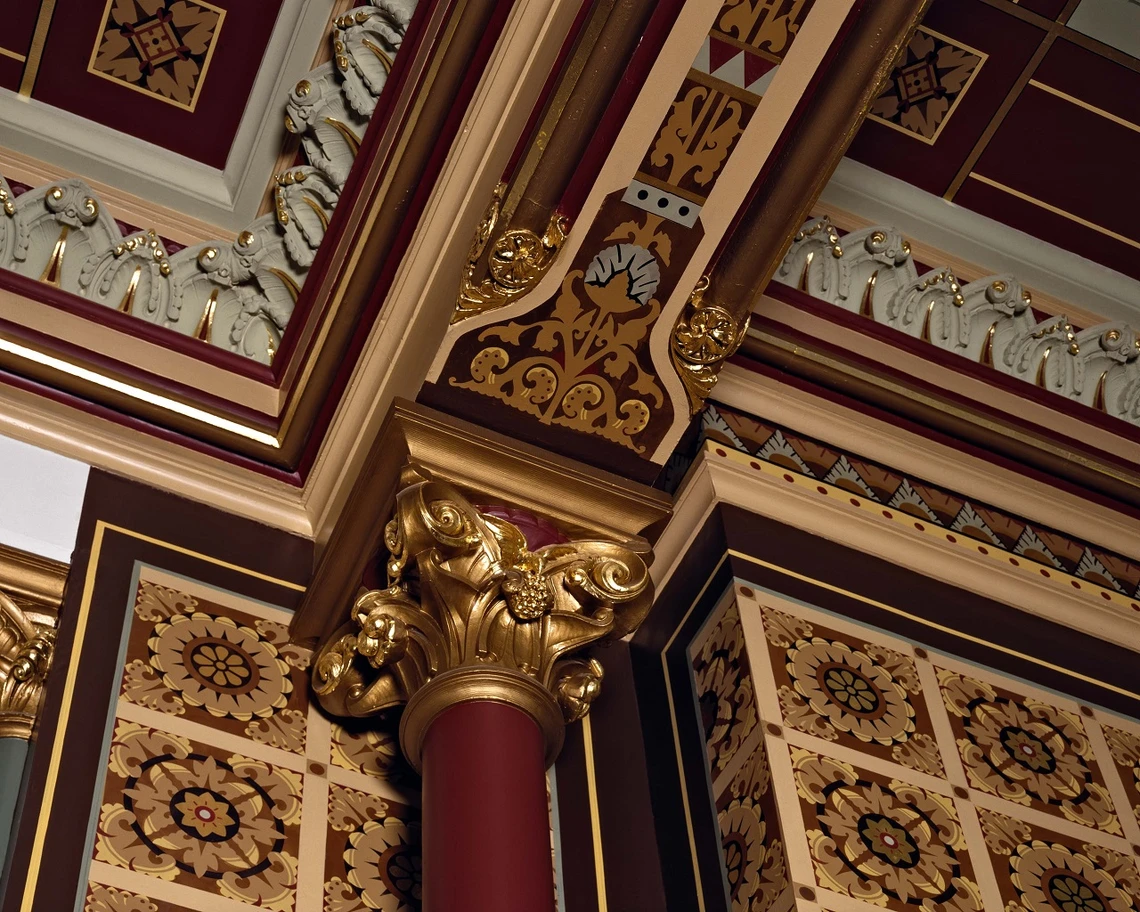 Close-up details of an elaboratively decorated room with painted brown, tan, and black floral patterns on the walls and ceiling and a gilding capital on top of a red-brown column.