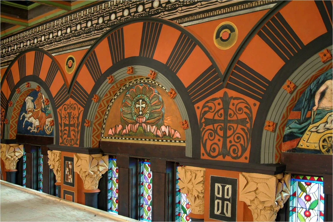 A wall near the ceiling of an historic church is decorated in a variety of decorative motifs and colors, including a border of black and tan geometric decorations above orange semi-circles surrounding beautifully painted scenes of chariots with horses and stylized crosses.