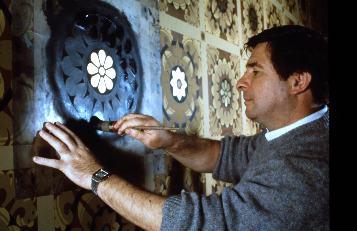 A man uses a paintbrush to paint a dark blue color onto a flower-shaped stencil attached to a wall; the colors of the finished painted decorations next to the piece he is stenciling are shades of brown and tan with dark blue accents.