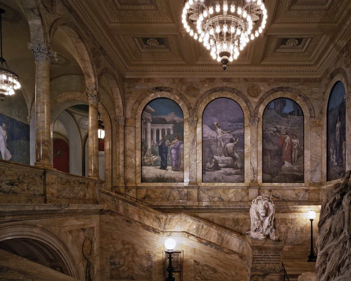 An elaborately decorated interior section of the Boston Public Library is filled with marble walls, columns, and a staircase; several large, painted murals decorate the walls, and a carved lion sits atop the staircase banister.