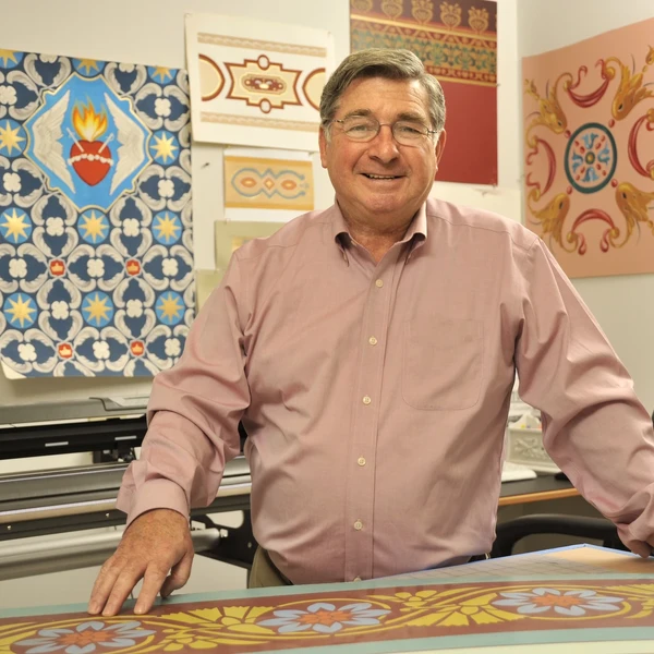 A man in a light pink shirt stands at a worktable in his studio with a variety of colorful decorative paintings on the wall behind him.