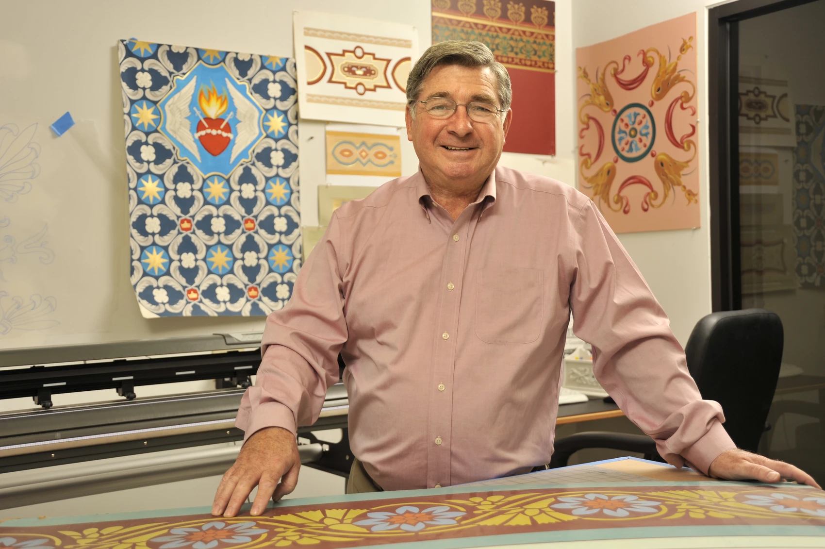 A man in a light pink shirt stands at a worktable in his studio with a variety of colorful decorative paintings on the wall behind him.