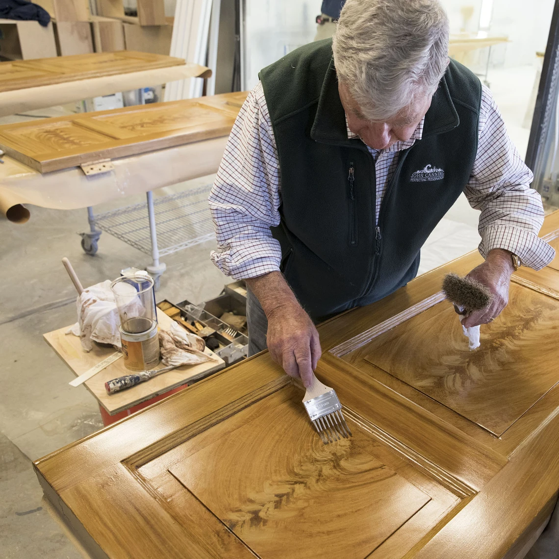 An older man in a workshop uses a comb-like brush to apply brown paint to a door on a worktable to imitate the patterns and colors of wood.