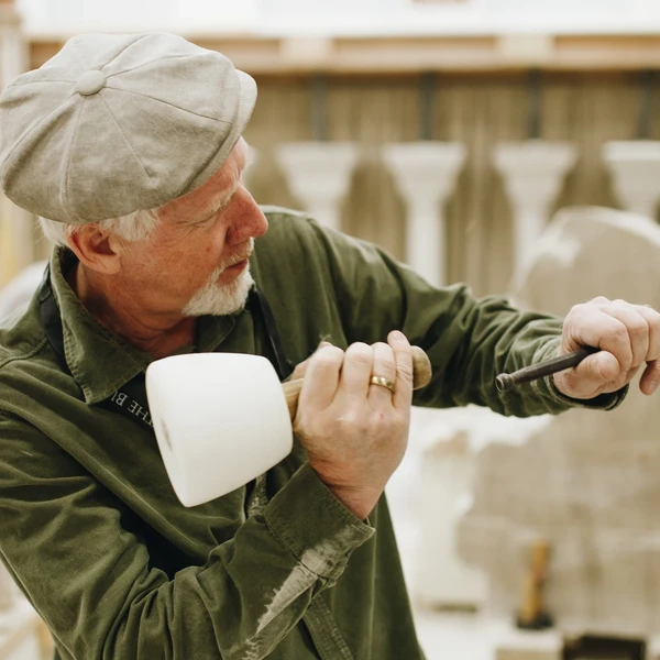A man in a studio uses a mallet and chisel to carve a piece of limestone.