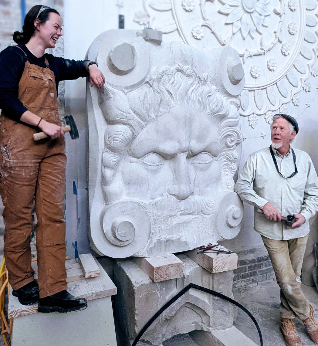 A young woman and a man in a stone carving studio are standing on either side of a large stone sculpture and smiling at each other. 