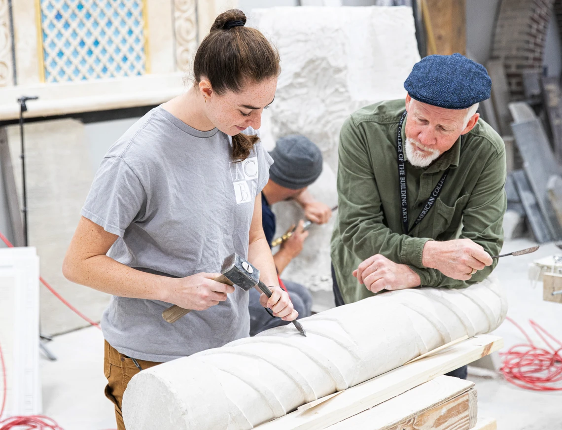 A man in a carver’s cap watches as a female college student carves an ornamental stone column with a mallet and chisel in the stone carving workshop at the American College of the Building Arts.