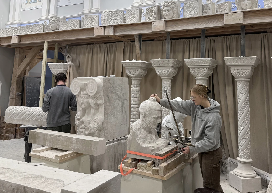 Two college students carve stone in a studio filled with pieces of stone and ornamental columns topped with carved capitals.