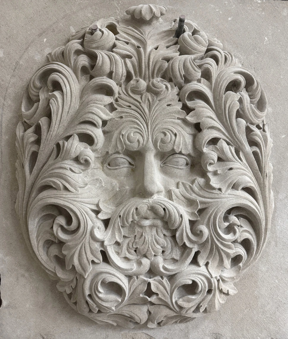 A fanciful oval-shaped decorative sculpture carved in limestone depicts the face of a man with a beard surrounded by intricate ornamental foliage.