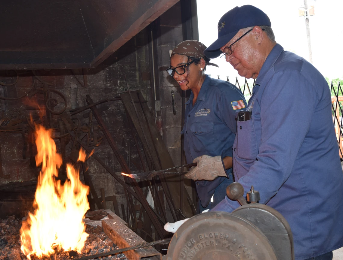 An older man and a young woman work together to heat a piece of metal in the orange flames of a coal-fired forge.