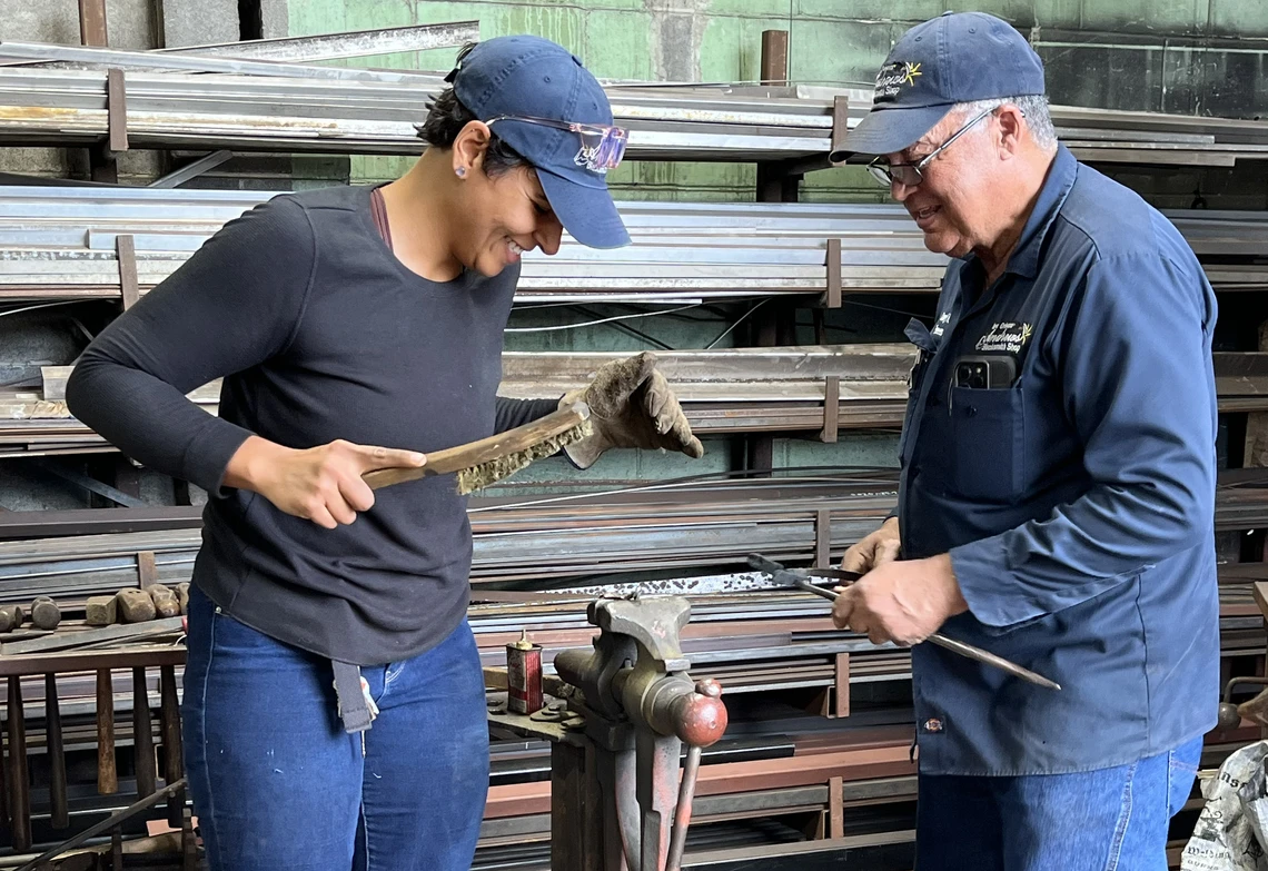 A young woman with a hard-wire brush and a man with long metal tongs work together to polish a hand-forged metal leaf clamped in a vise.
