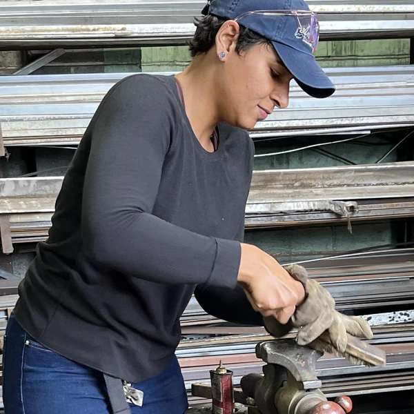 A young woman in a blacksmith’s shop uses a hard-wire brush to polish a piece of metal clamped in a vise; rows of shelves stacked with long metal rods line the wall behind her.