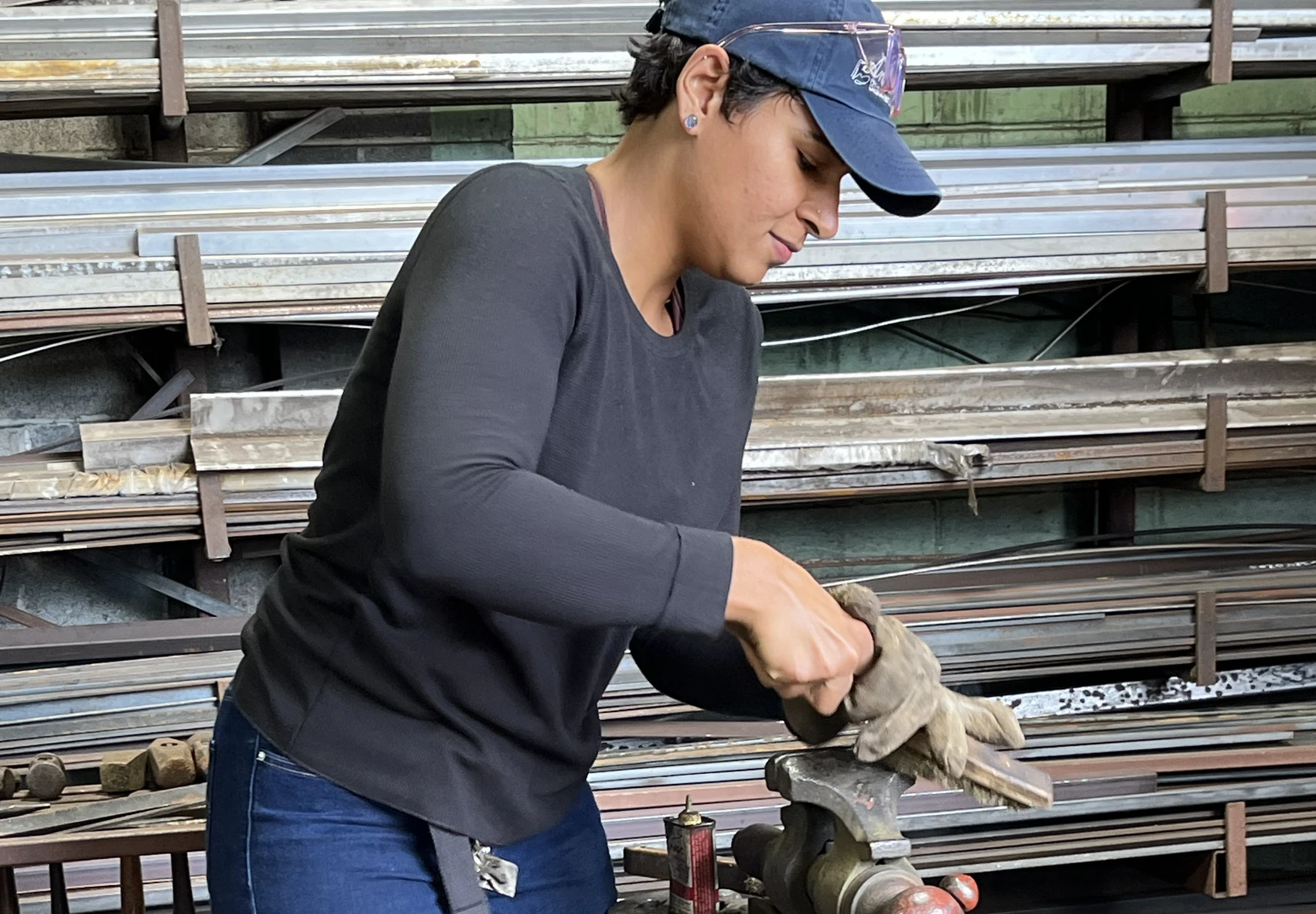 A young woman in a blacksmith’s shop uses a hard-wire brush to polish a piece of metal clamped in a vise; rows of shelves stacked with long metal rods line the wall behind her.