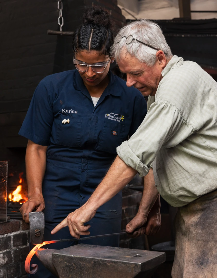A man shows a young woman how to use a hammer to shape hot metal on an anvil in a blacksmith’s shop.