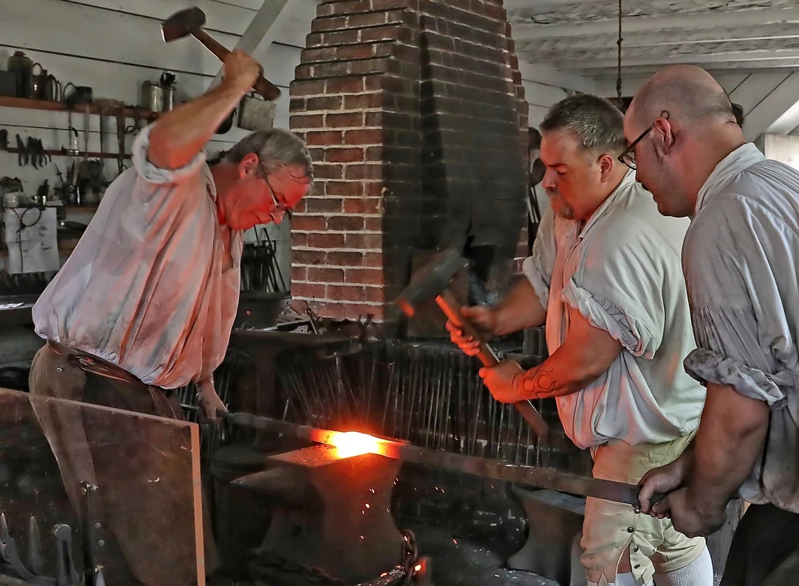 Three men in a blacksmith’s shop use big hammers to beat a hot piece of metal on an anvil.