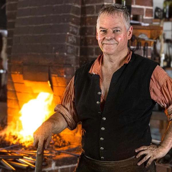 A man in a blacksmith’s shop stands at his anvil with a hammer in his hand and the fire of the forge behind him.