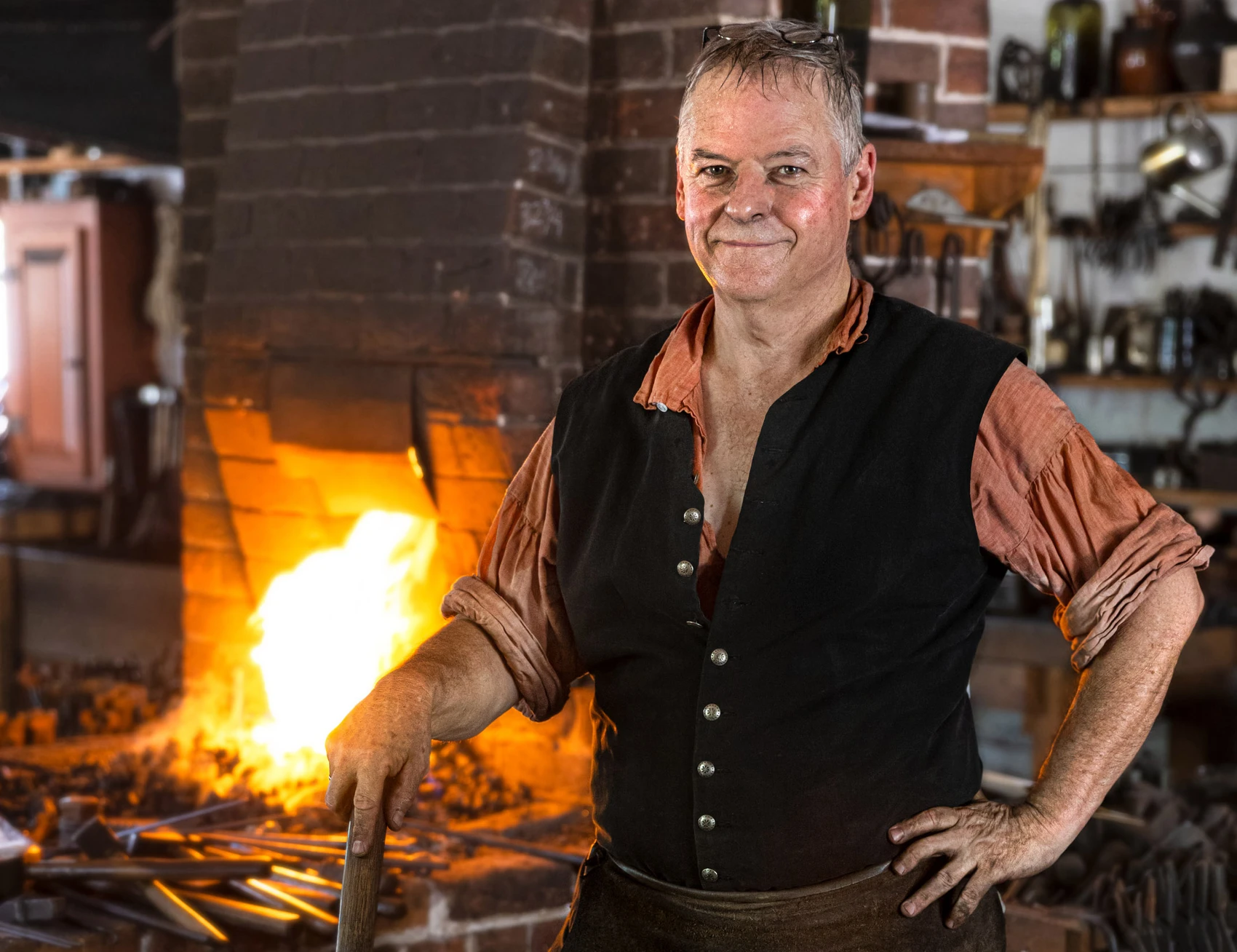A man in a blacksmith’s shop stands at his anvil with a hammer in his hand and the fire of the forge behind him.
