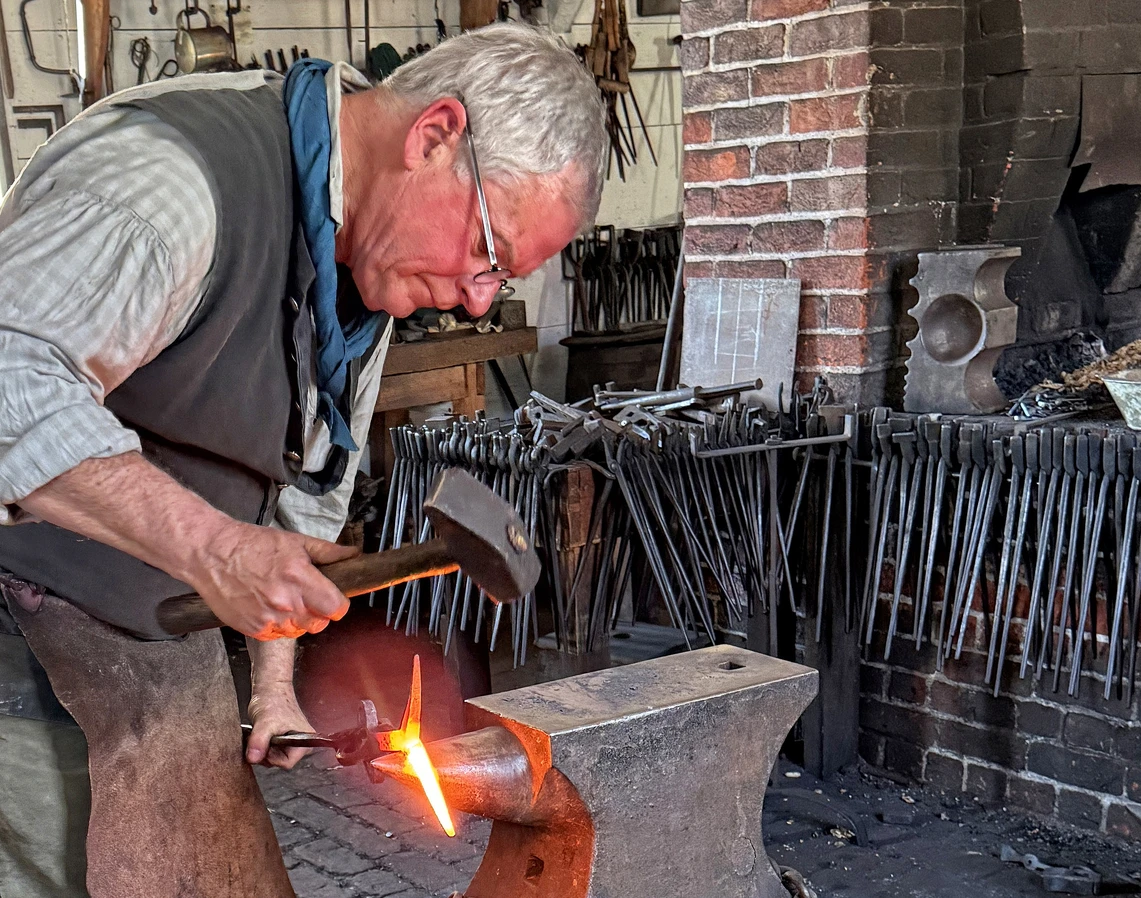 A man in a blacksmith’s shop uses a hammer to shape hot metal on an anvil.