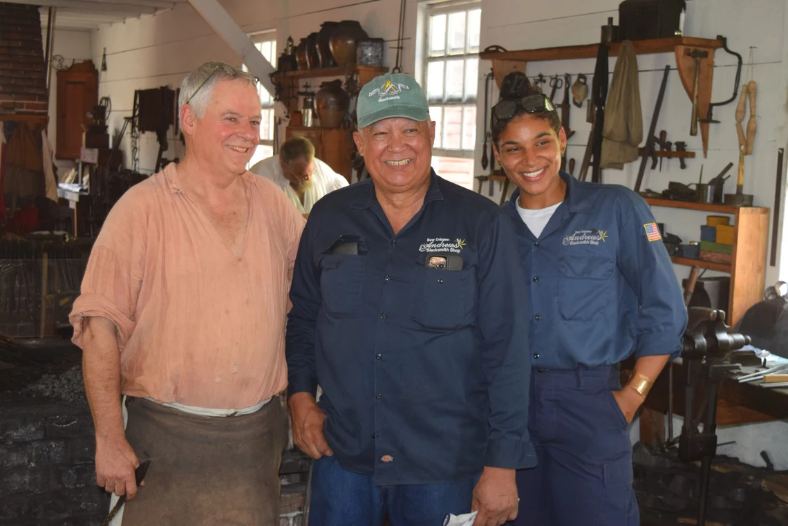 Two men and a woman with big smiles of their faces pose in a blacksmith’s shop.