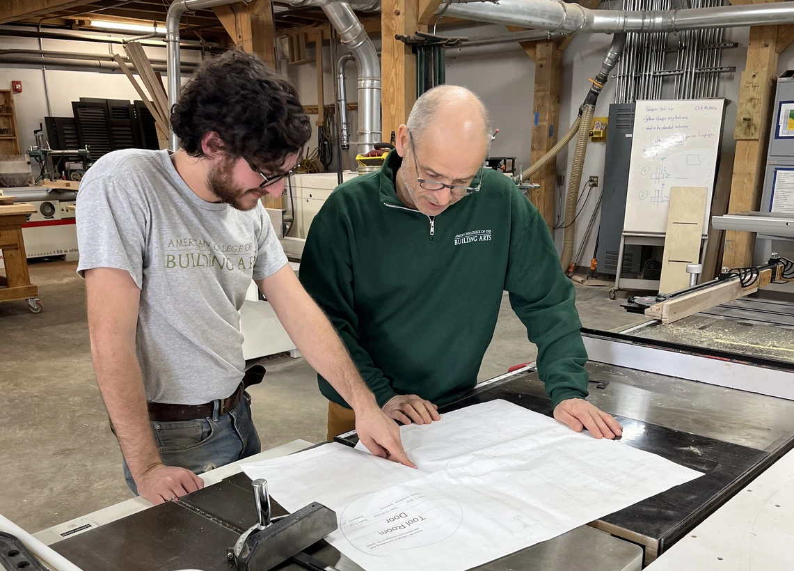 A male carpentry teacher and a young male college student look at architectural drawings together in a carpentry workshop.