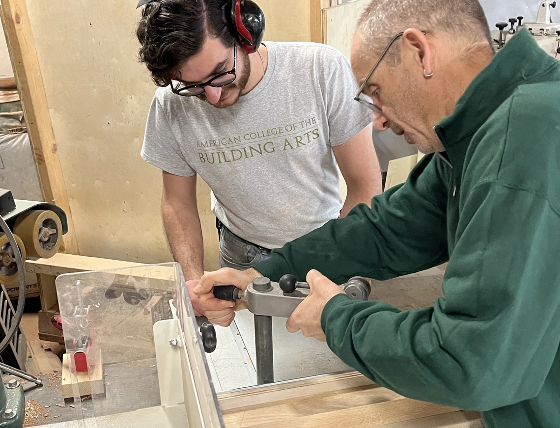 A male carpentry teacher shows a young male college student how to clamp a piece of wood to a workbench in a carpentry workshop.