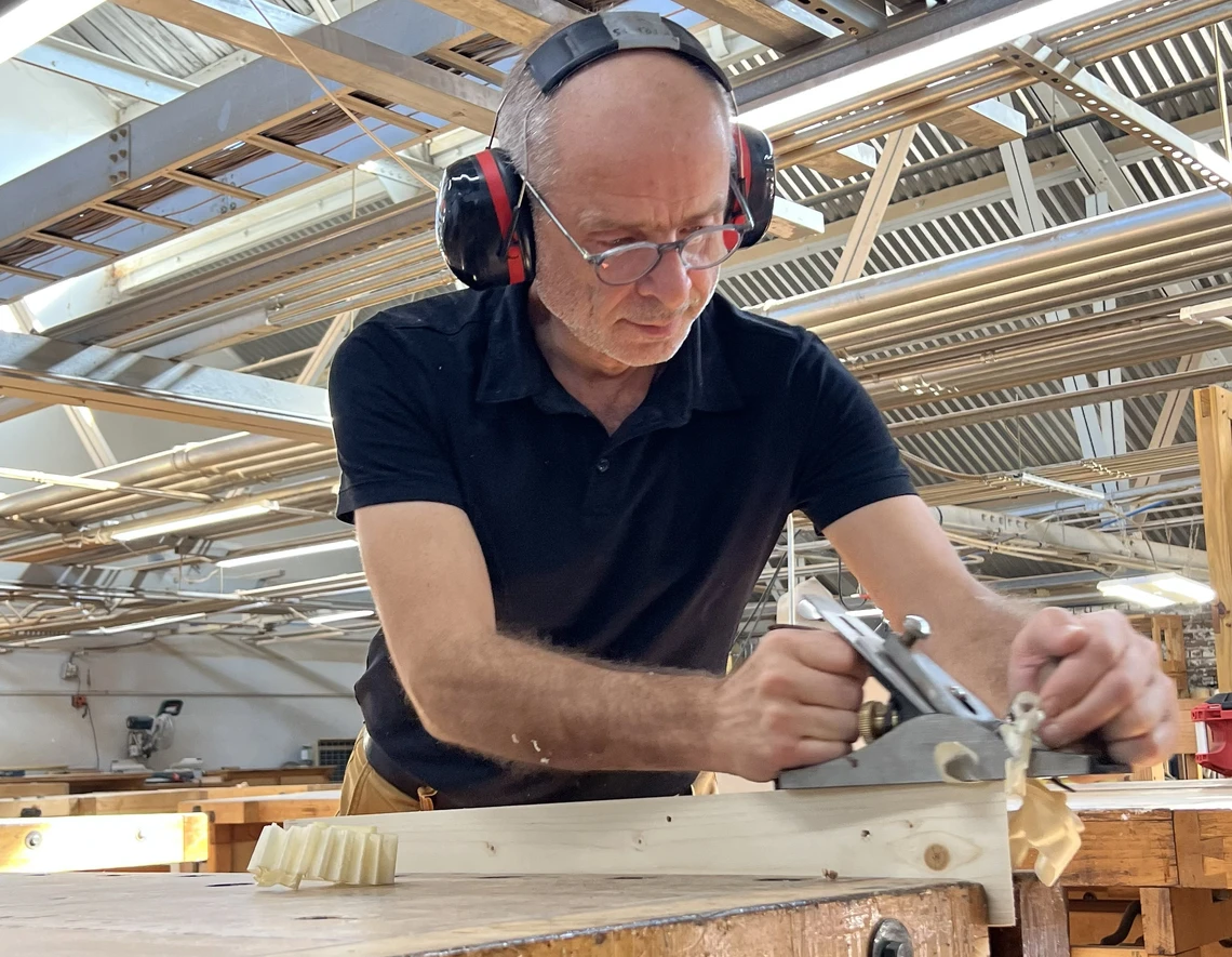 A man uses a metal plane to smooth a piece of wood on a worktable in a carpentry workshop.