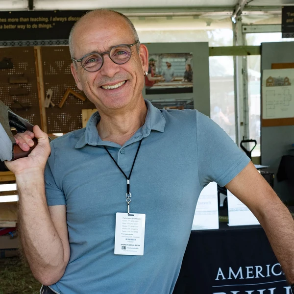 A man holding a woodworking plane poses outside in a tent with a smile on his face.