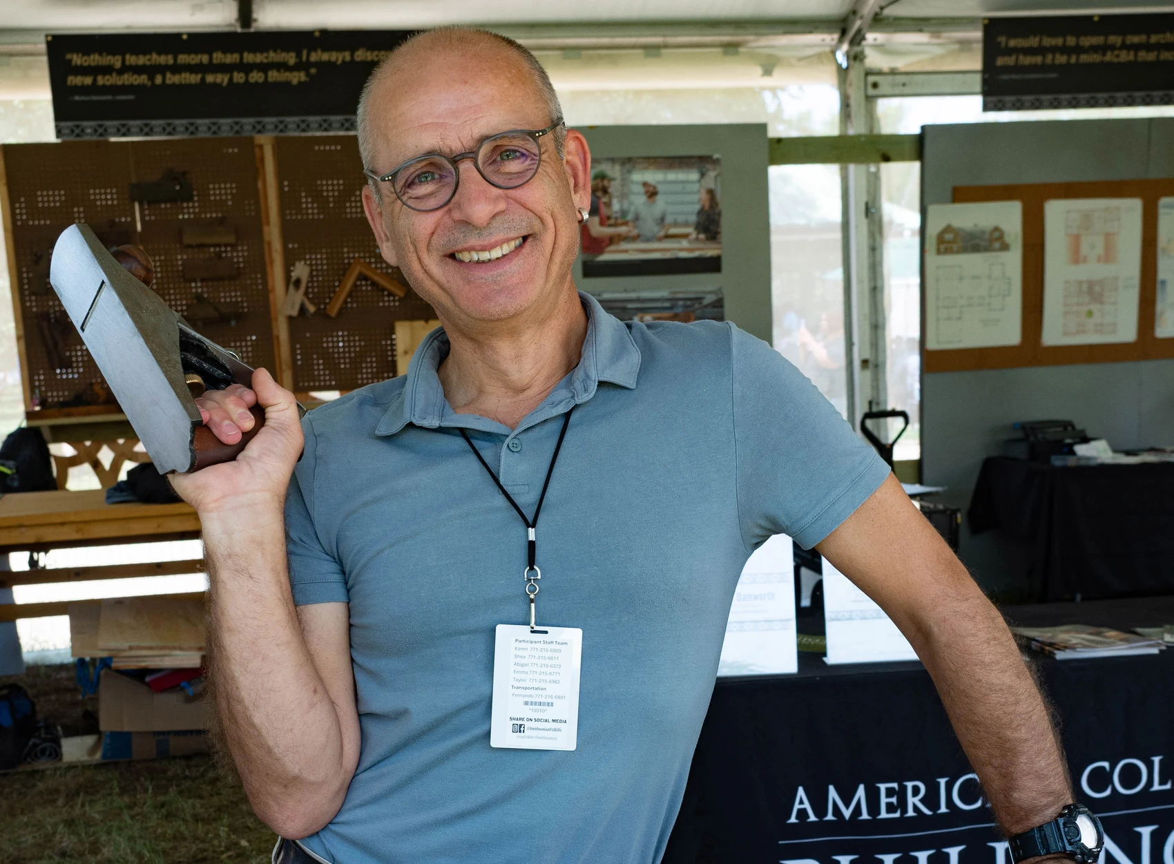 A man holding a woodworking plane poses outside in a tent with a smile on his face.
