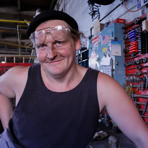 A man with glasses on top of his head and a smile on his face poses in a blacksmith’s shop.