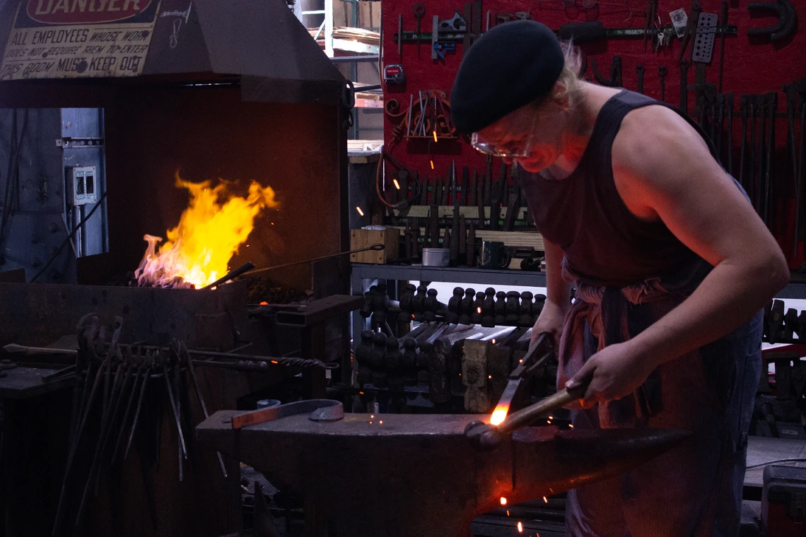 A man shapes a hot piece of metal on an anvil inside a blacksmith’s shop; a wall with tools and a forge with the orange flames of a fire is behind him.