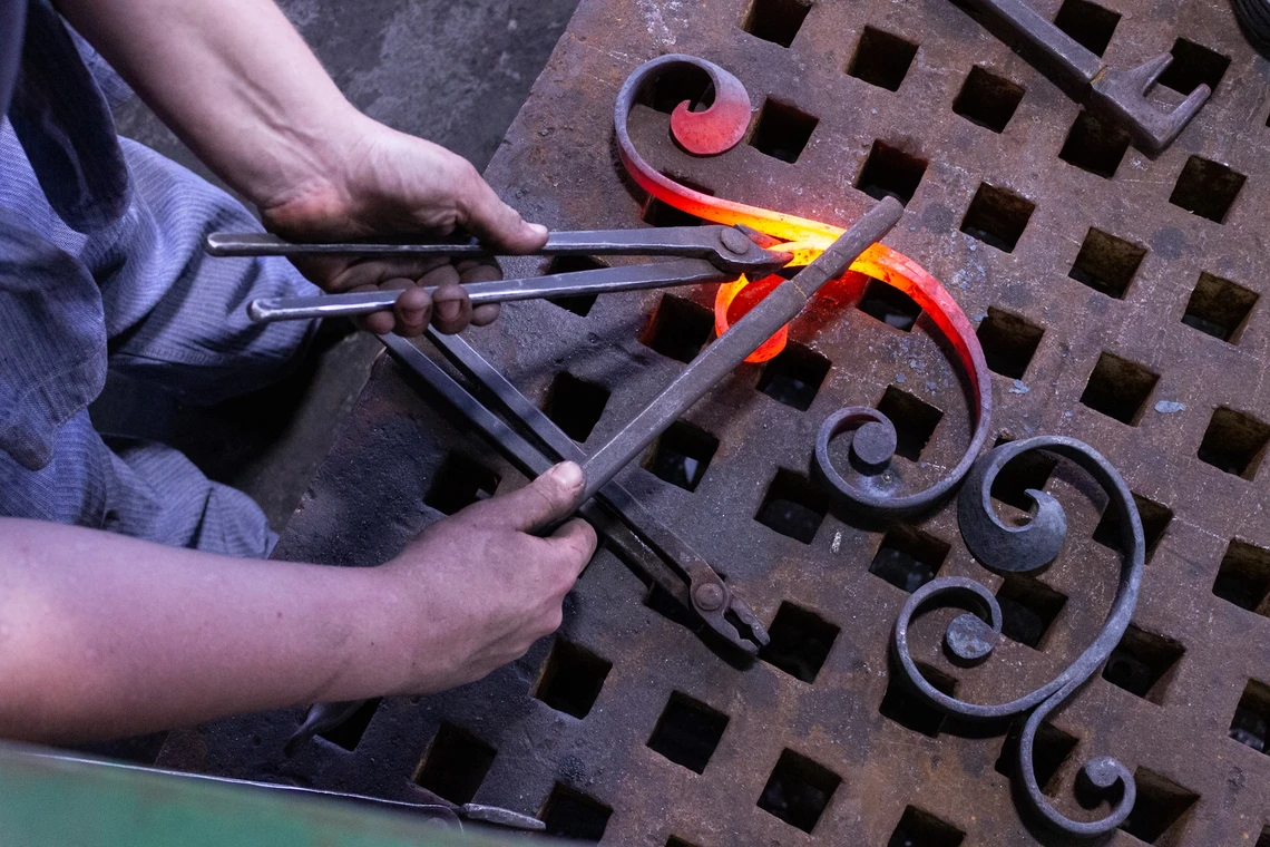 This image shows a close-up of a man’s hands holding metal tongs to shape a red-hot piece of metal into a scroll on a worktable.