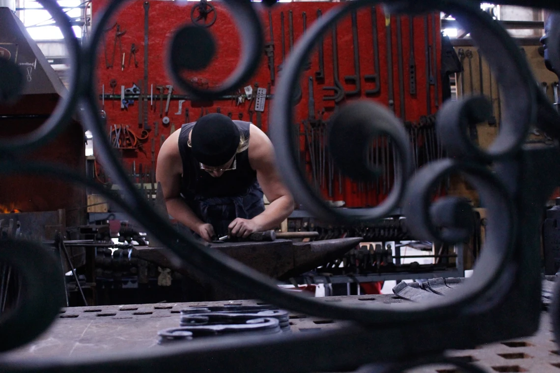 A man is bending over to craft a piece metal on an anvil in a blacksmith’s shop; a large metal scroll is in the foreground.