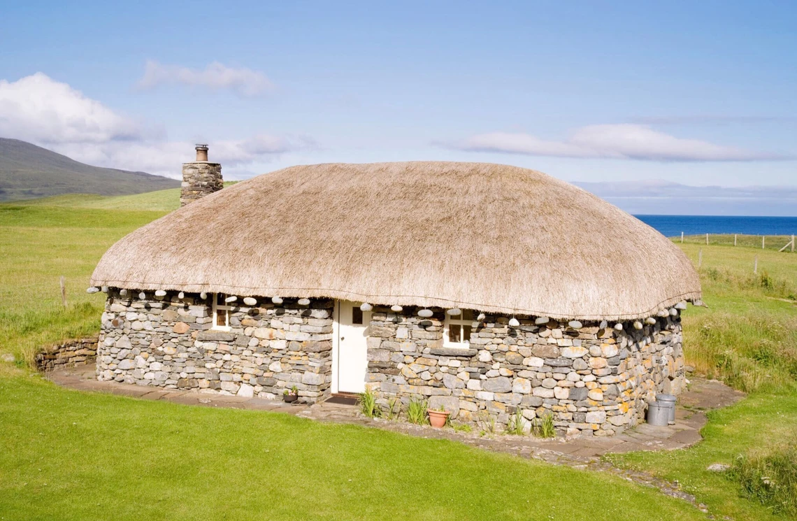 A small drystone house with a thatched roof sits in a green field with a view of the ocean behind it.