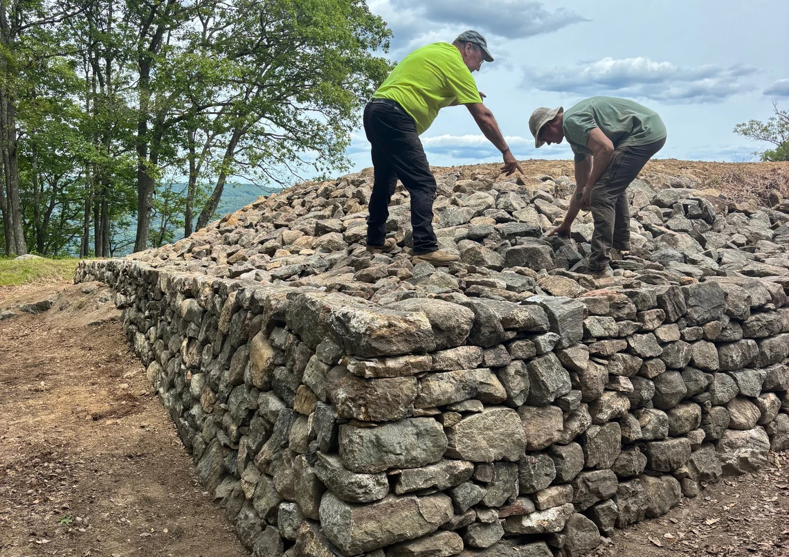 Two men place stones on top of a large mound of grey stones that forms the remains of an old historic stone fort in need of repair.