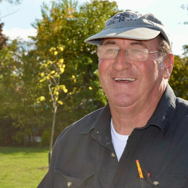 A man with glasses and a blue hat and shirt stands outside on a green lawn with a smile on his face.