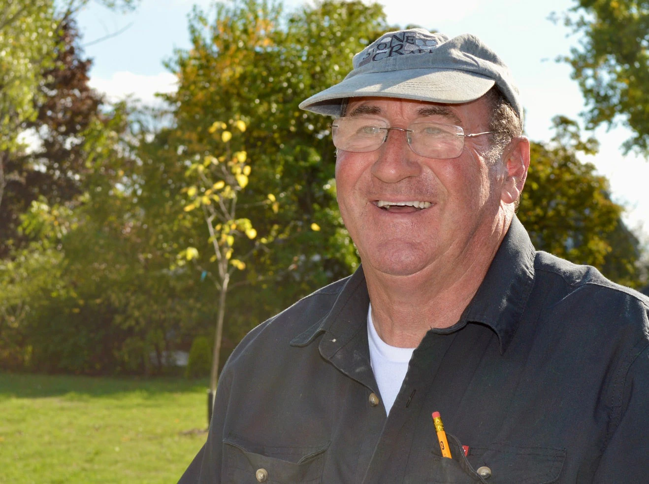 A man with glasses and a blue hat and shirt stands outside on a green lawn with a smile on his face.