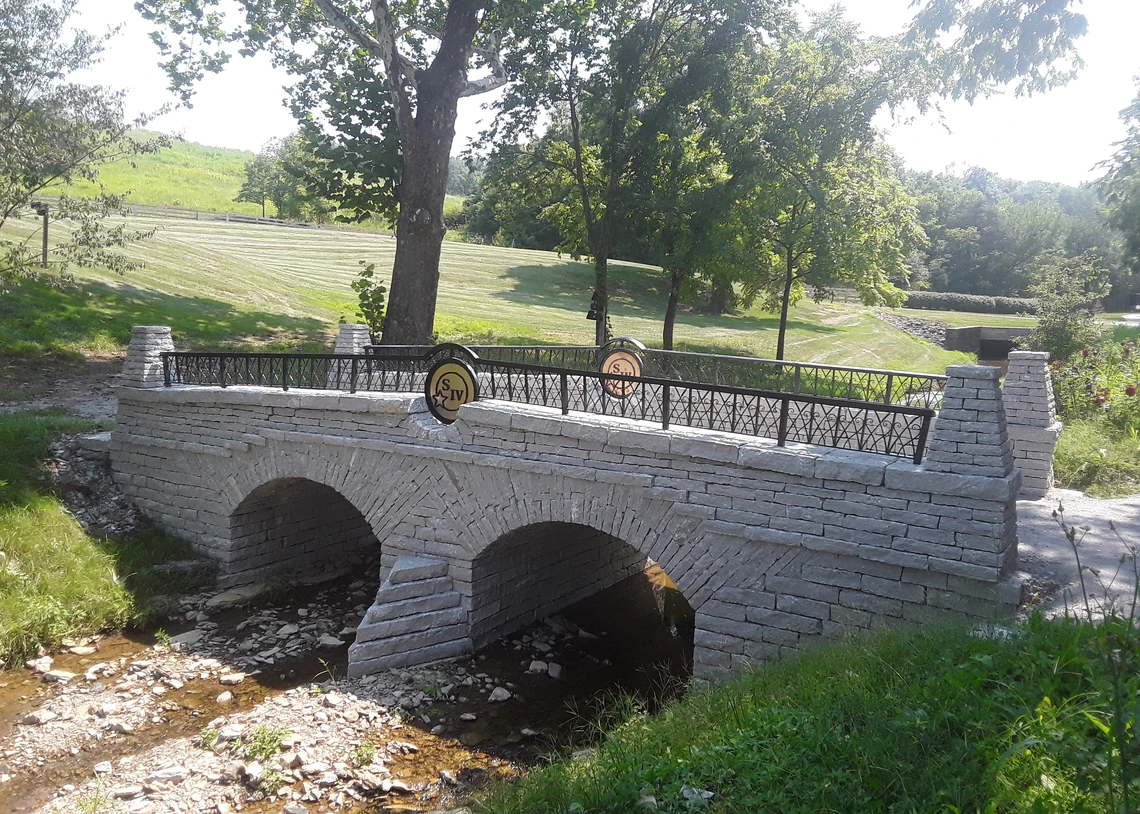 A double-arched drystone bridge crosses over a creek in the midst of green fields.    