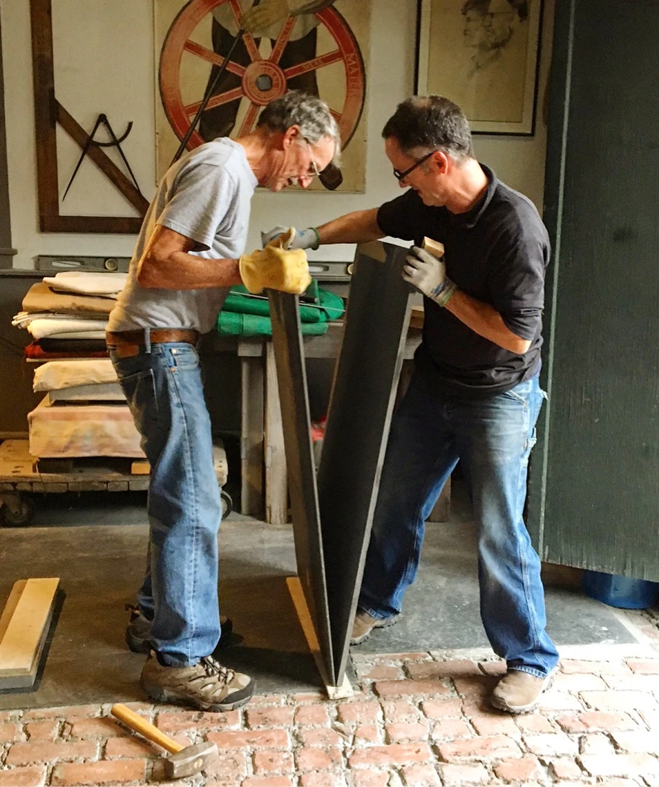 Two men in a stone carving shop work together to split a large vertical piece of slate.