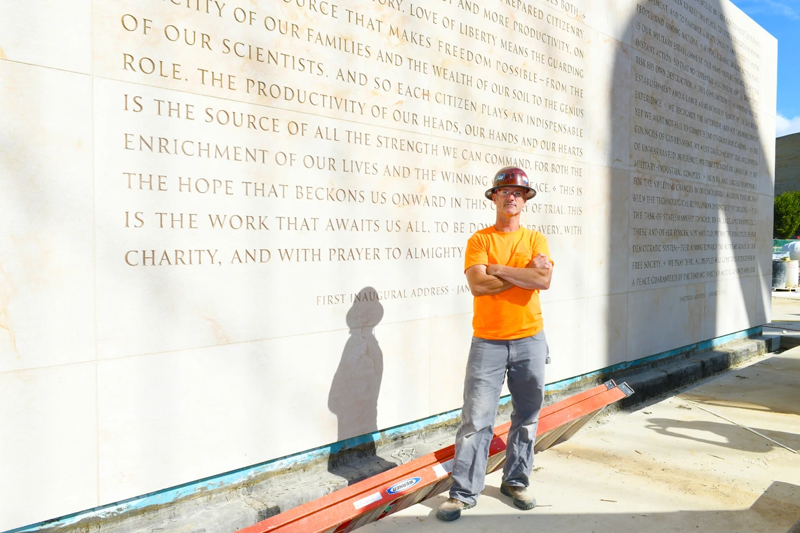 A man in a hard hat and orange t-shirt stands outside in front of a large stone wall filled with carved inscriptions.