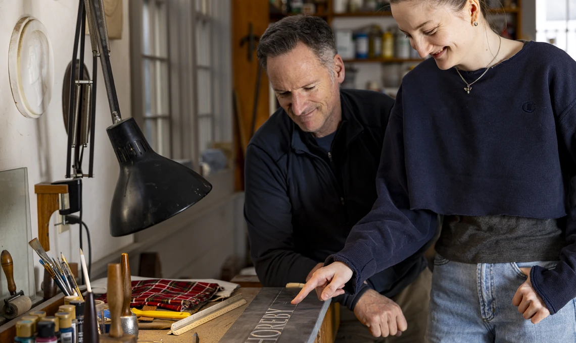 A young woman in a stone carving studio shows the man next to her a piece of stone with hand-carved letters.
