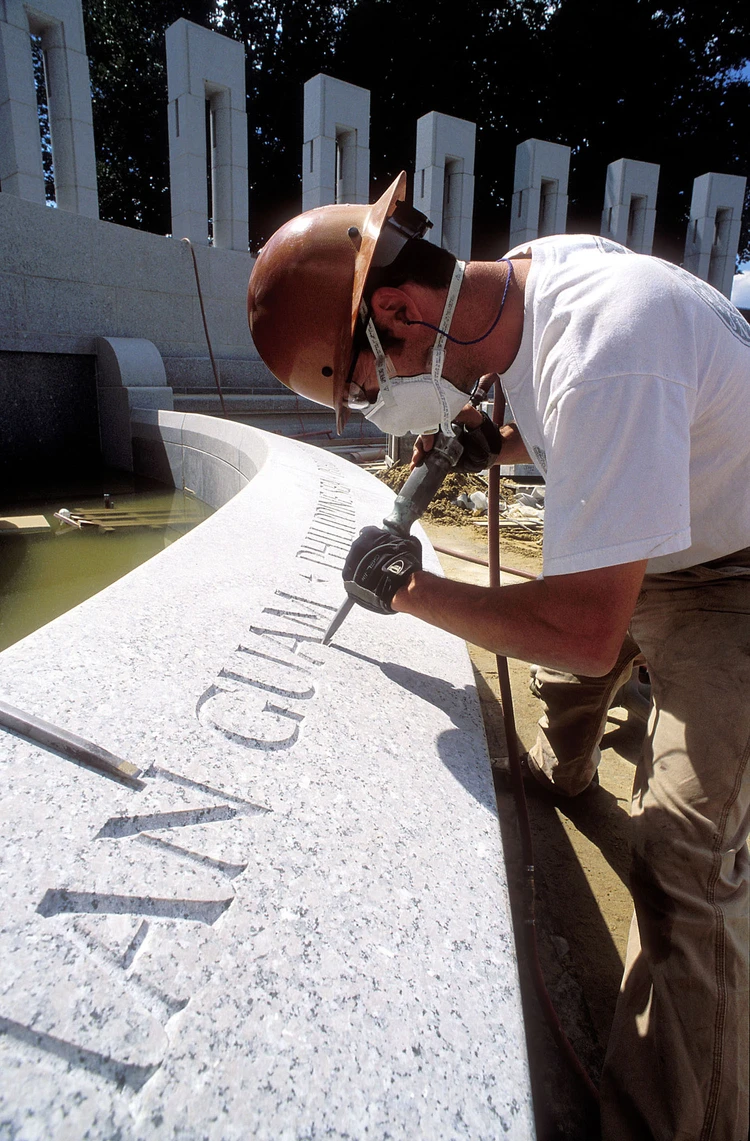 A man in a hard hat working outside at the World War II Memorial uses a pneumatic hammer and chisel to carve letters in granite.