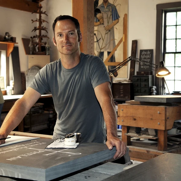 A man in a stone carving workshop poses at a workbench with a piece of slate with hand-carved inscriptions; two other men are carving pieces of stone at worktables behind him.