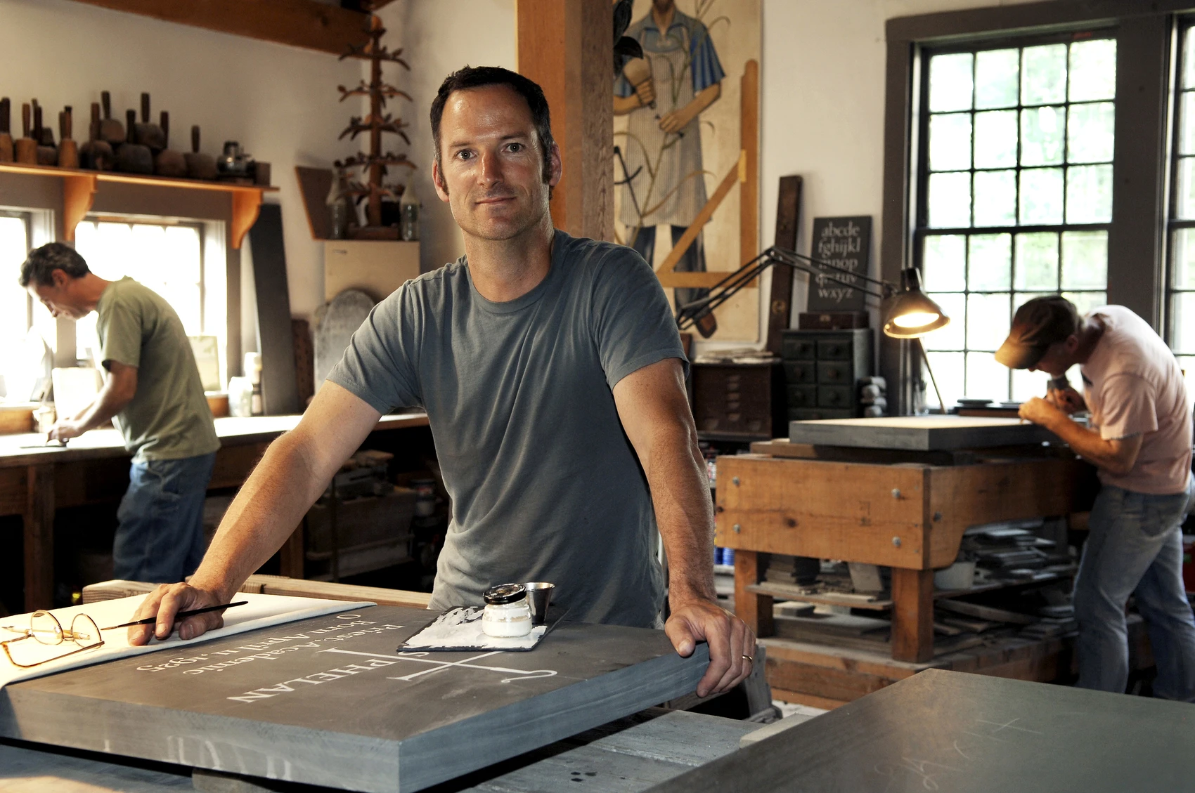 A man in a stone carving workshop poses at a workbench with a piece of slate with hand-carved inscriptions; two other men are carving pieces of stone at worktables behind him.