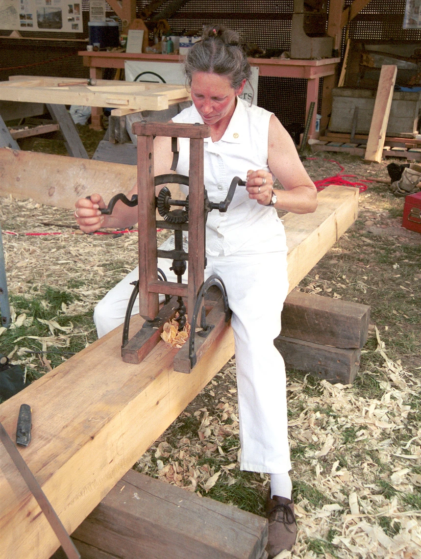 A woman straddling a large squared-off wooden beam uses a hand-powered drill to cut a hole in the beam.