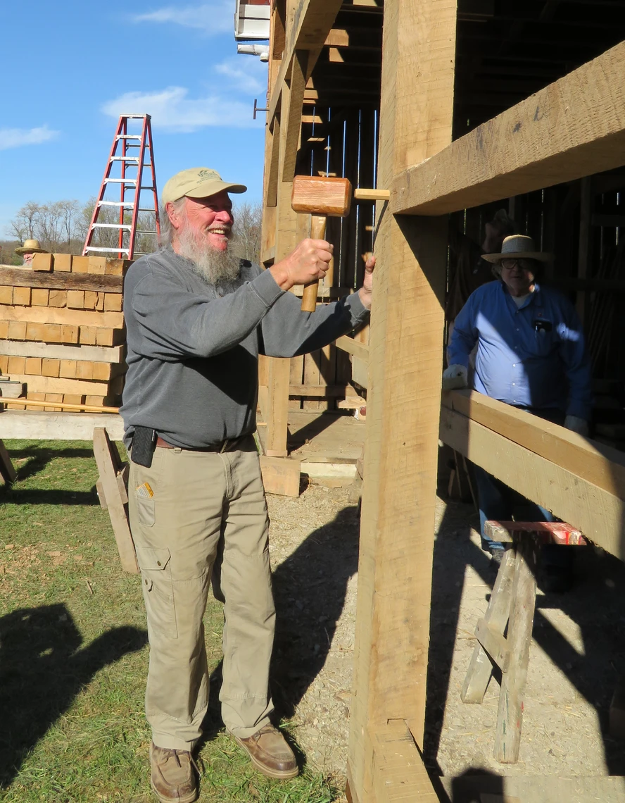 A man with a white beard uses a wooden mallet to drive a wooden peg into the vertical beam of a timber-frame structure