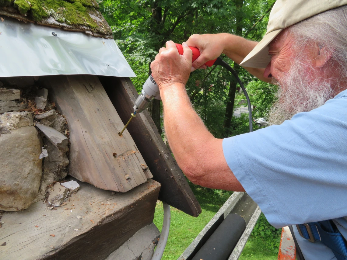 A man with a white beard works in scaffolding to drill a hole in an old wooden beam of a roof.