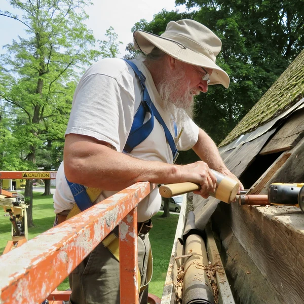 A man with a white beard standing in scaffolding uses a wooden mallet to repair the roof of an old wooden building.