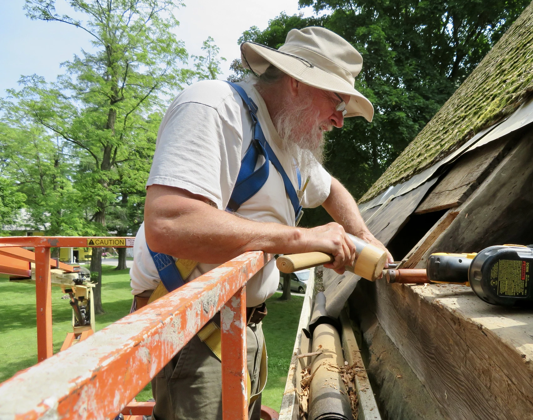 A man with a white beard standing in scaffolding uses a wooden mallet to repair the roof of an old wooden building.