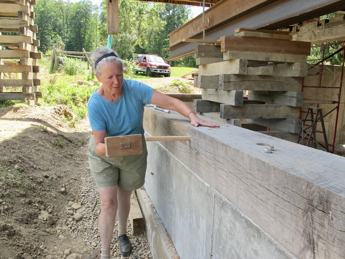 A woman uses a large wooden mallet to pound a wooden peg into the side of a wooden beam.